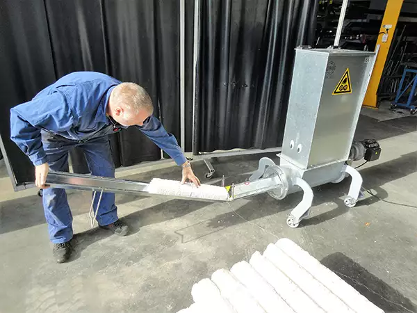 A man stands bent over as he pulls compacted Styrofoam tubes from the Styropactor Nano. The Styropactor Nano has four curved legs and a sticker of two eyes on the front of its metal cuboid shell.
